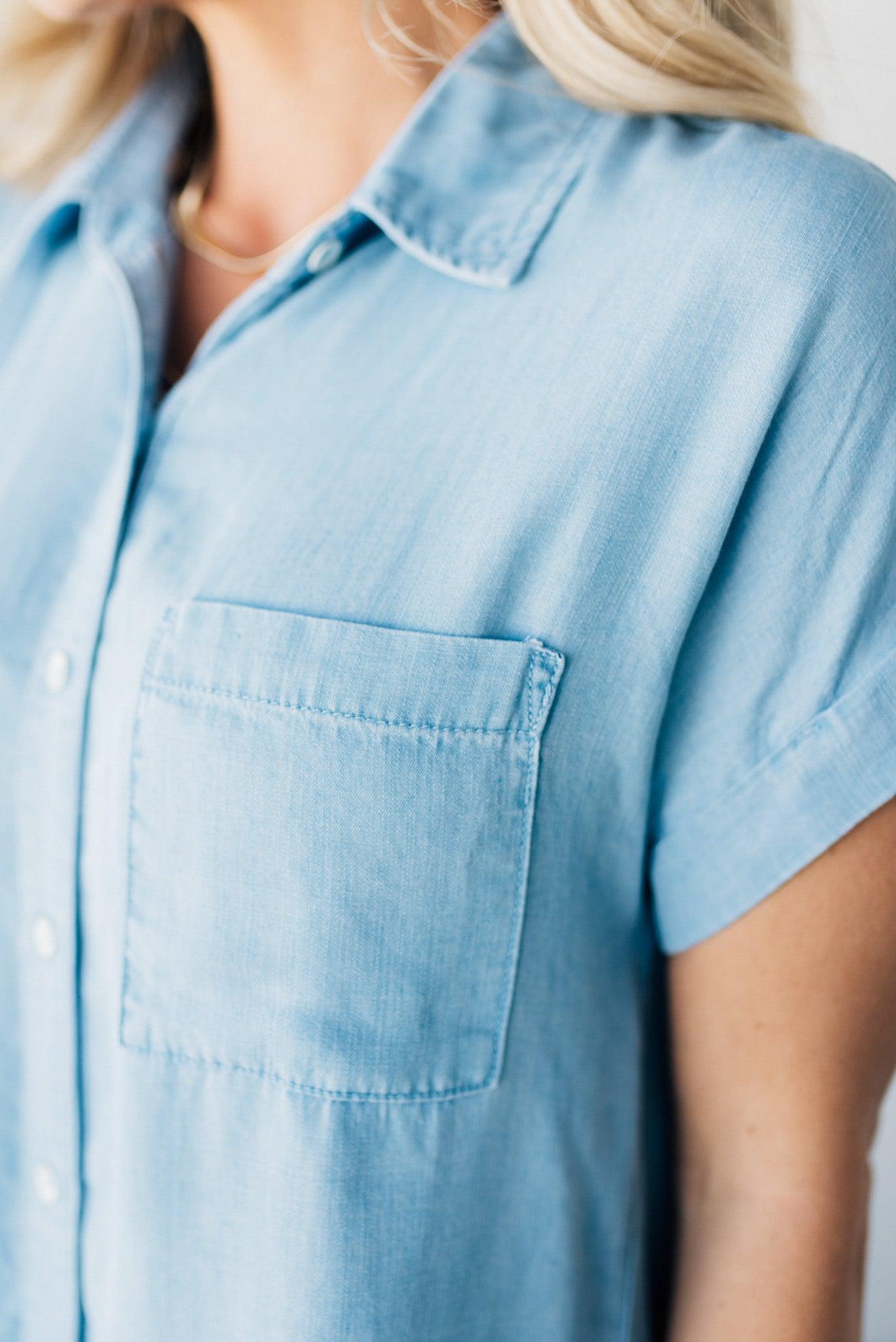 A woman smiling and wearing a blue chambray button front top with a patch chest pocket, dolman shoulders, and short sleeves rolled up to the elbows.