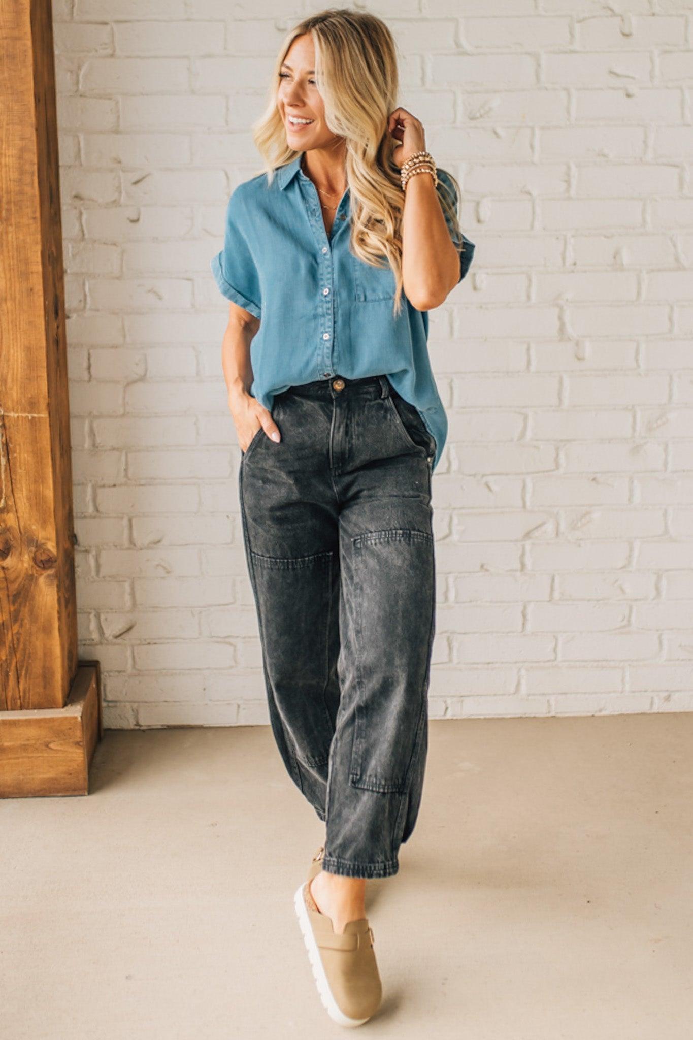 A woman smiling and wearing a blue chambray button front top with a patch chest pocket, dolman shoulders, and short sleeves rolled up to the elbows.