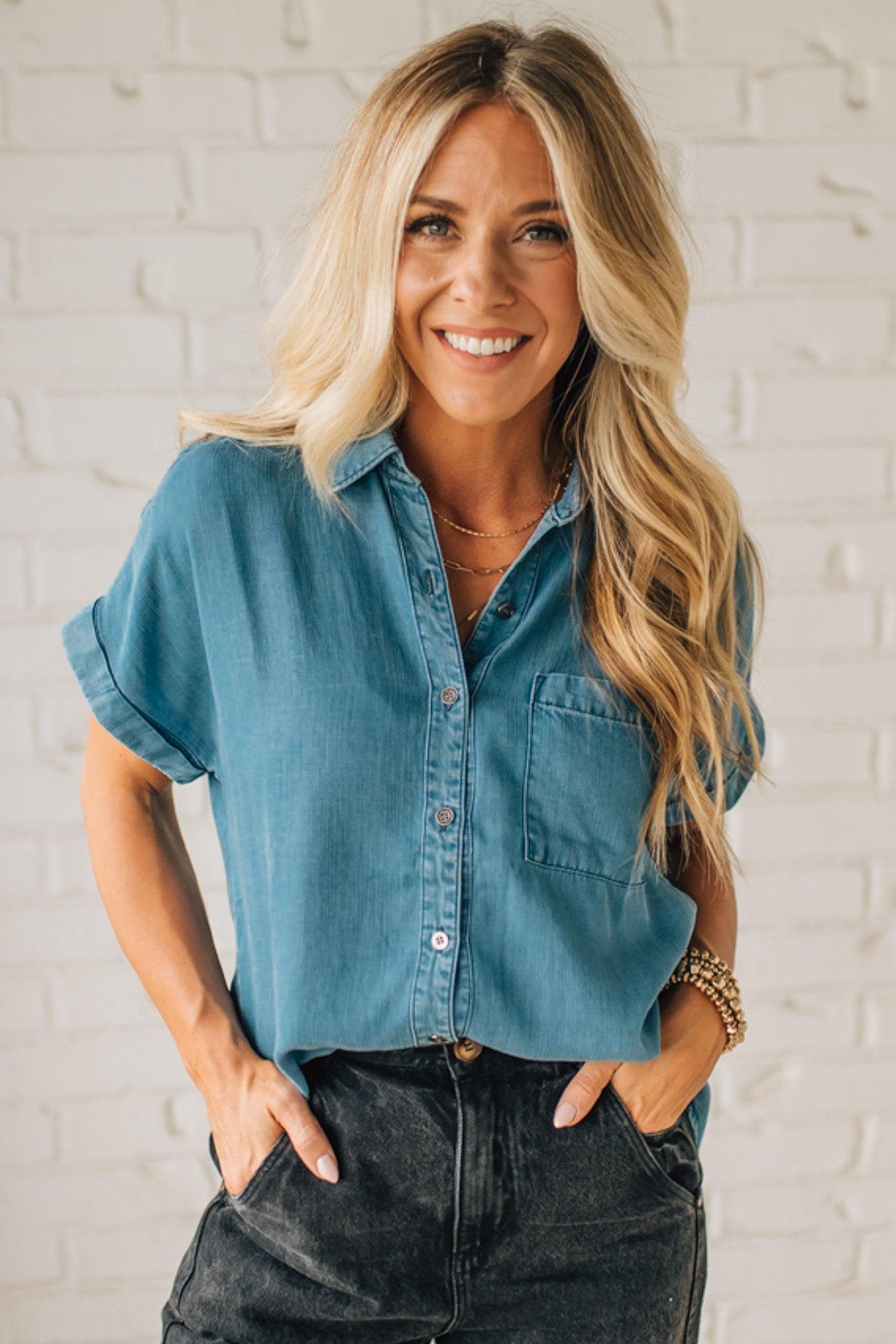 Woman wearing a blue denim shirt and black pants against a white brick wall.