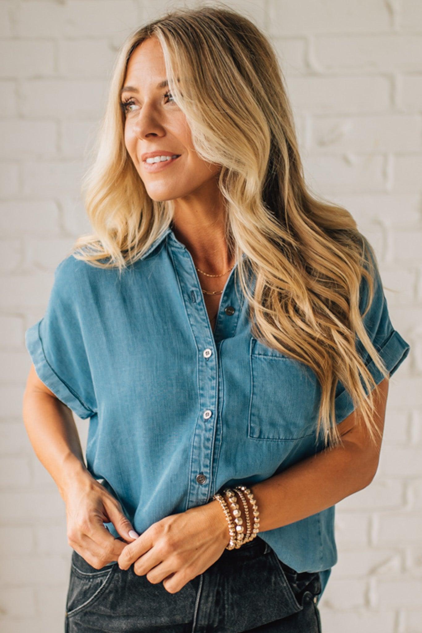 A woman smiling and wearing a blue chambray button front top with a patch chest pocket, dolman shoulders, and short sleeves rolled up to the elbows.