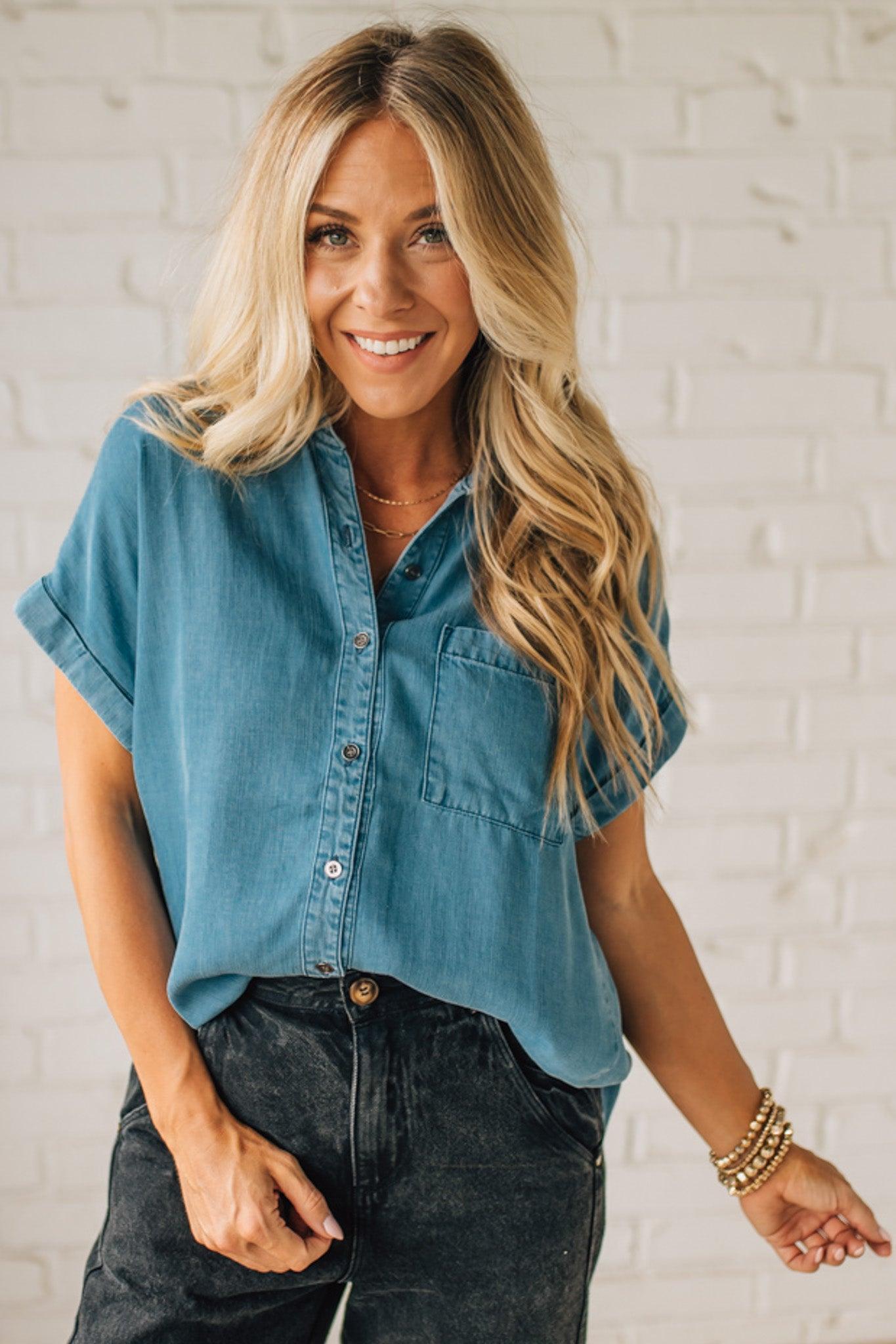 A woman smiling and wearing a blue chambray button front top with a patch chest pocket, dolman shoulders, and short sleeves rolled up to the elbows.