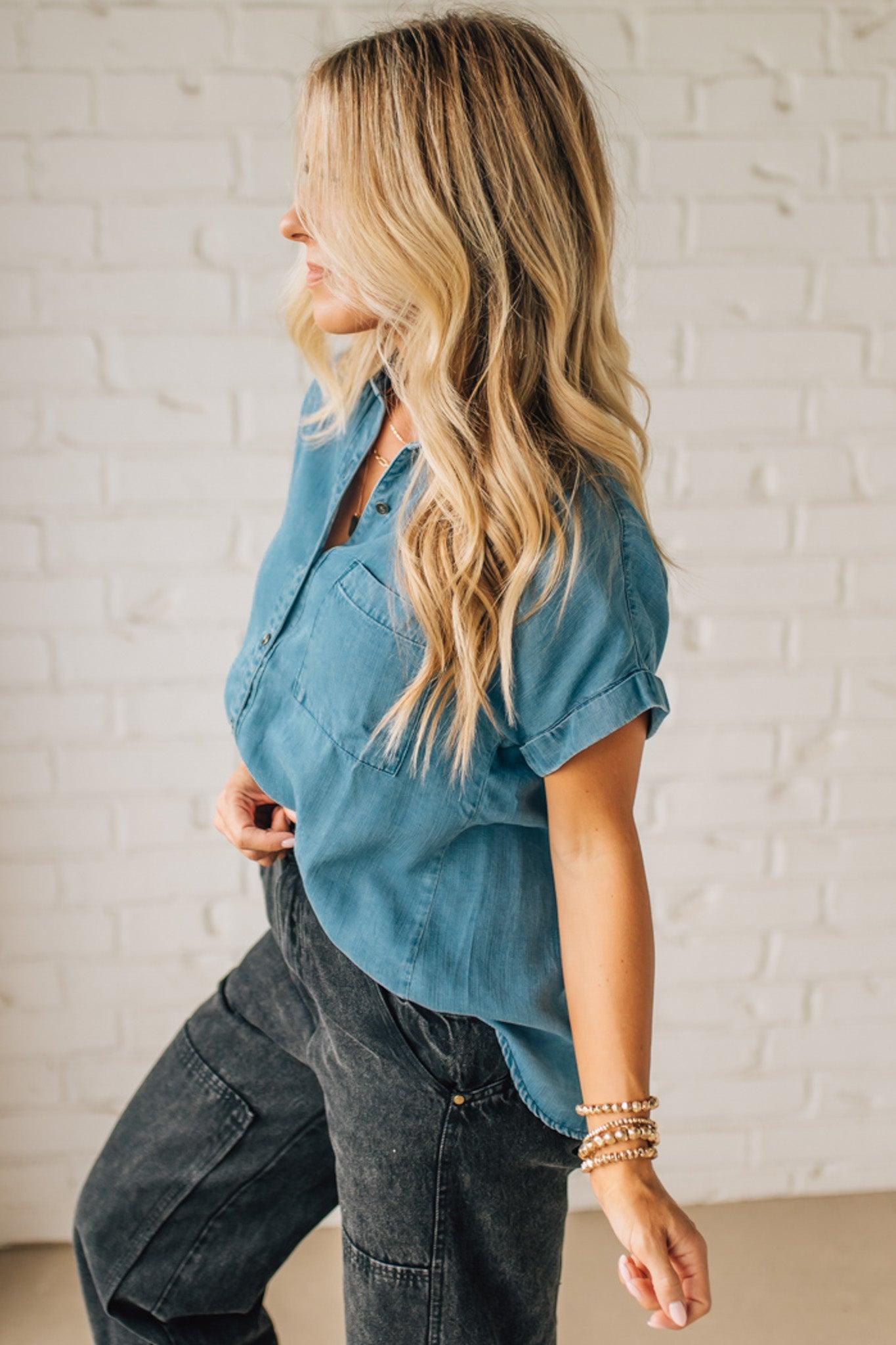 Woman wearing a blue denim shirt and dark jeans against a white brick wall.