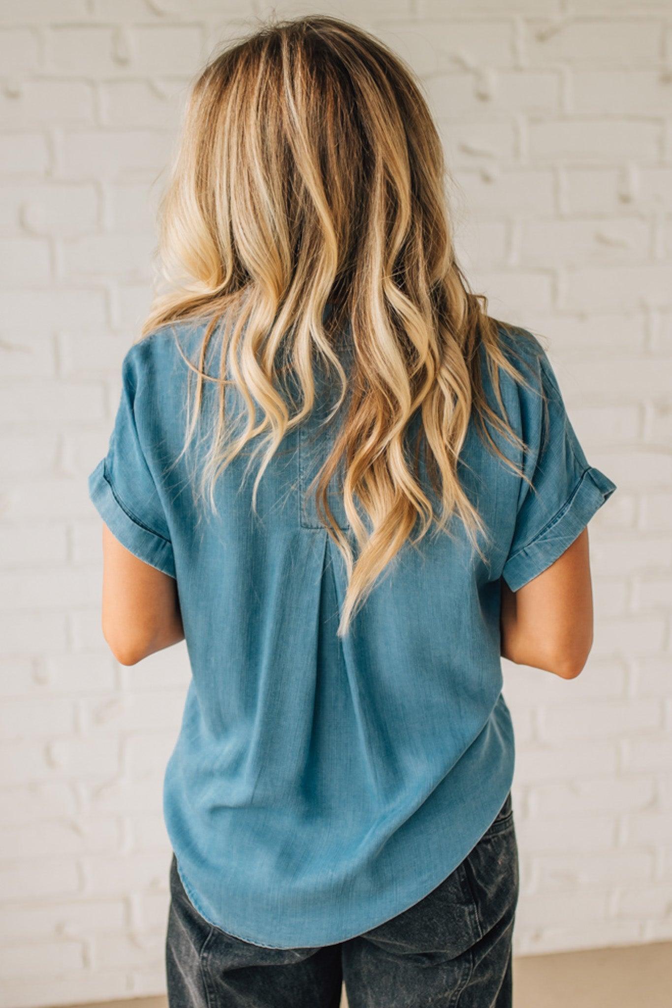 A woman smiling and wearing a blue chambray button front top with a patch chest pocket, dolman shoulders, and short sleeves rolled up to the elbows.