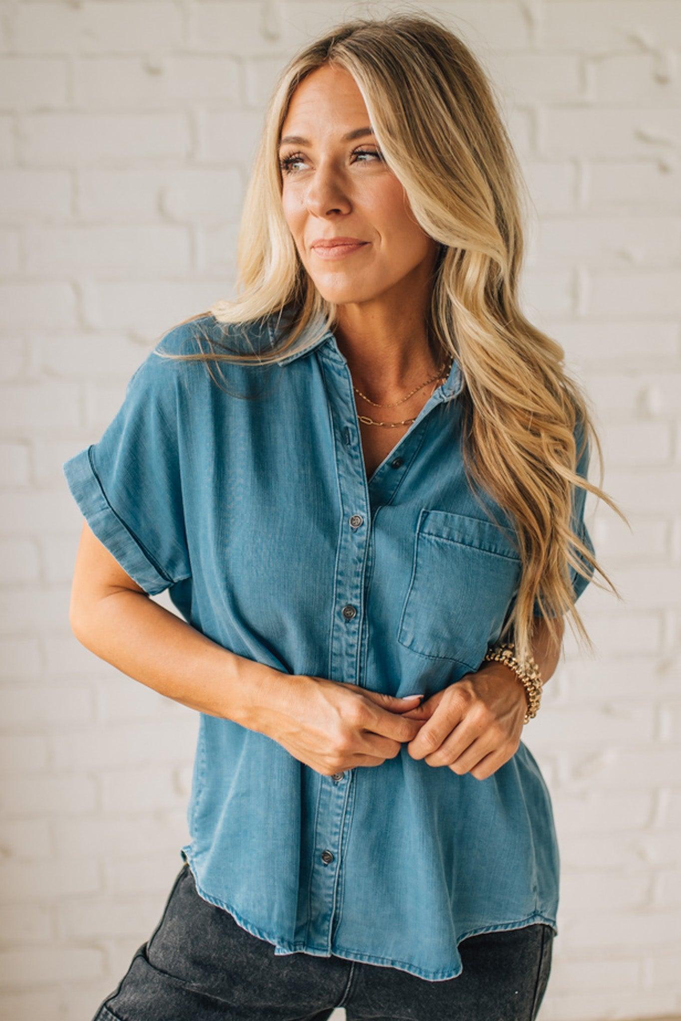 A woman smiling and wearing a blue chambray button front top with a patch chest pocket, dolman shoulders, and short sleeves rolled up to the elbows.