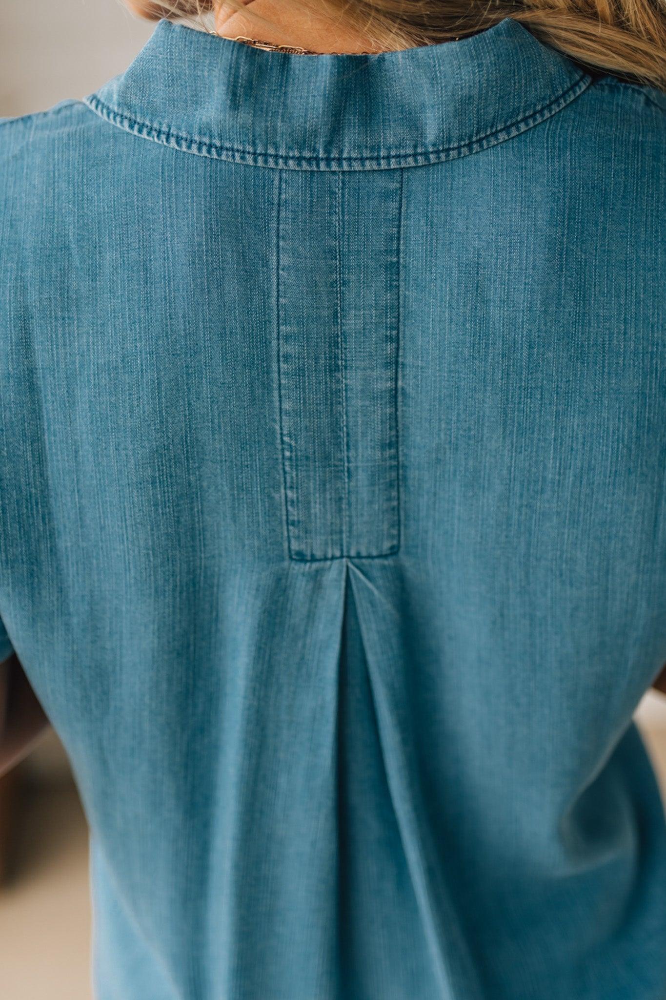 A woman smiling and wearing a blue chambray button front top with a patch chest pocket, dolman shoulders, and short sleeves rolled up to the elbows.