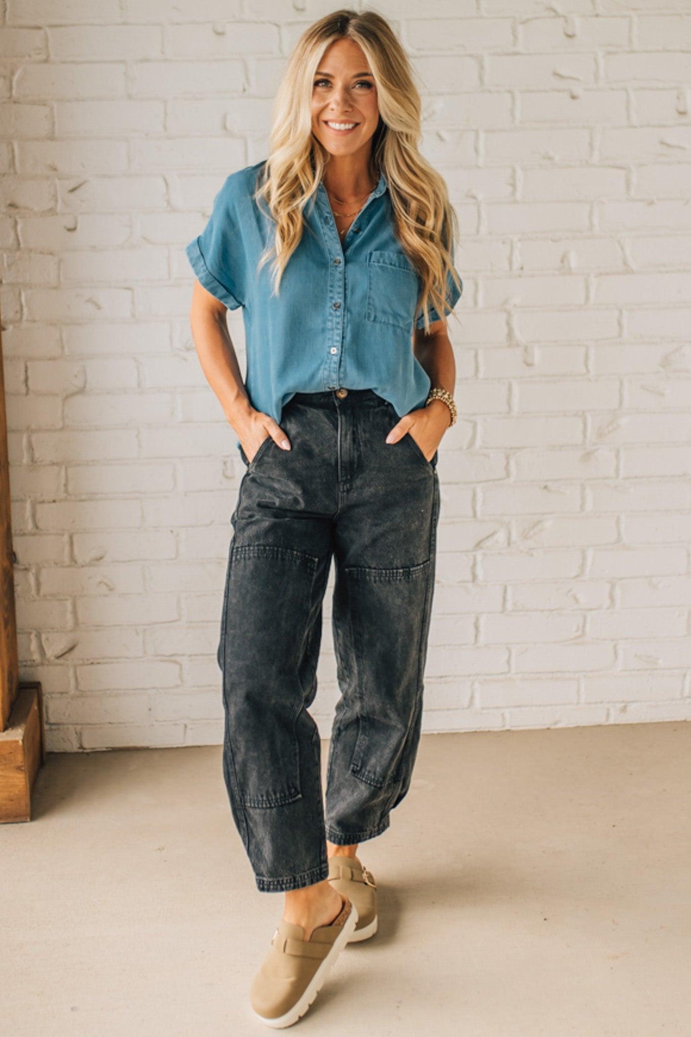 A woman smiling and wearing a blue chambray button front top with a patch chest pocket, dolman shoulders, and short sleeves rolled up to the elbows.