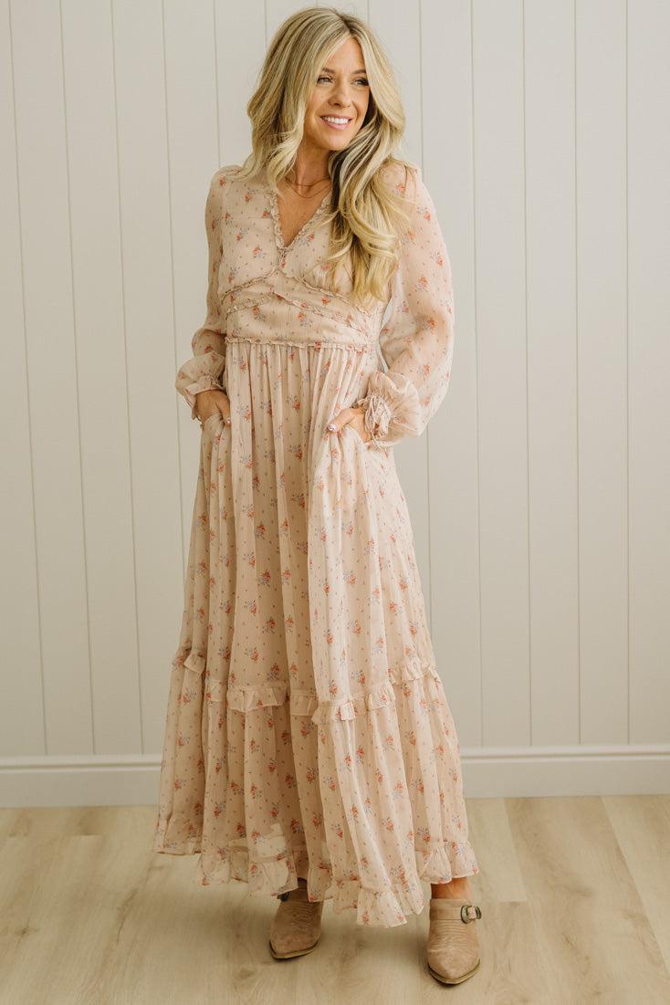 Woman wearing a long, light-colored floral dress standing against a white paneled wall.