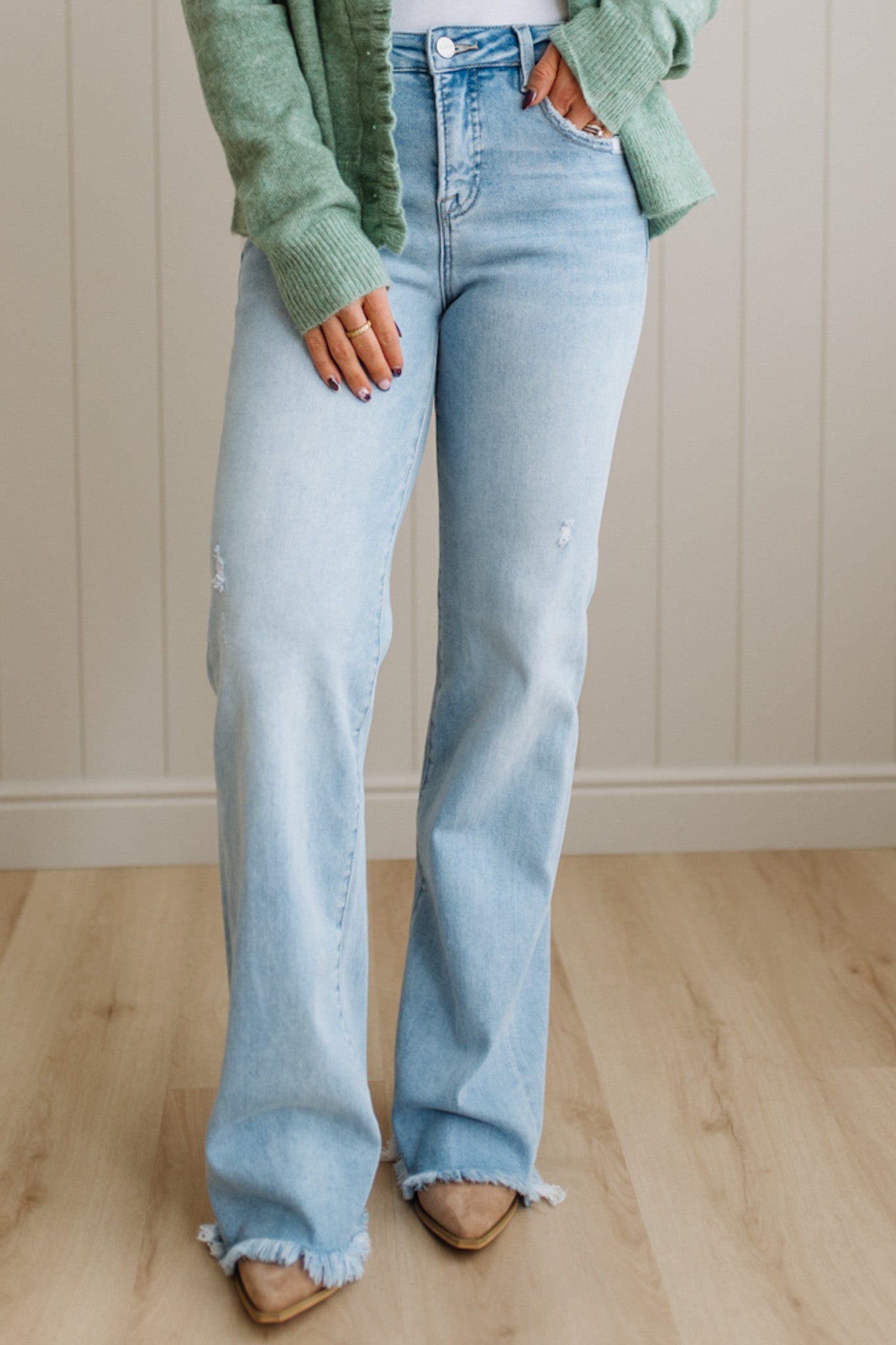 Person wearing light blue flared jeans and a green sweater against a neutral wall.