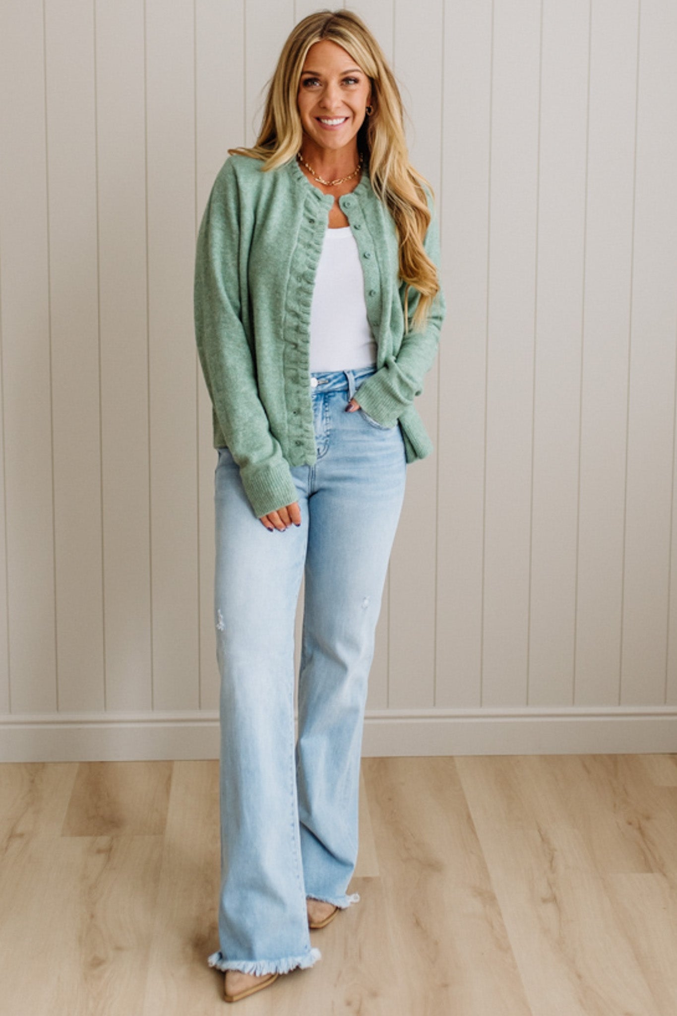 Woman wearing a green jacket, white top, and light blue jeans standing against a beige wall.