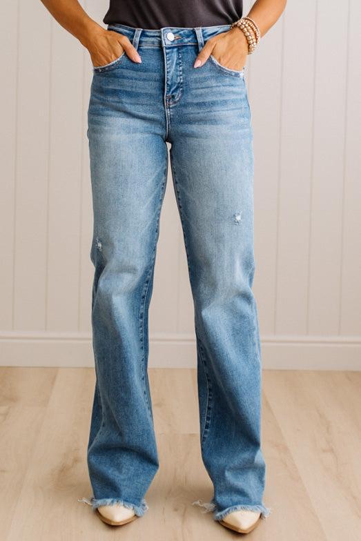 A person wearing light blue high-waisted flare jeans with a raw frayed hem, standing in front of a white paneled wall and light wood floor.