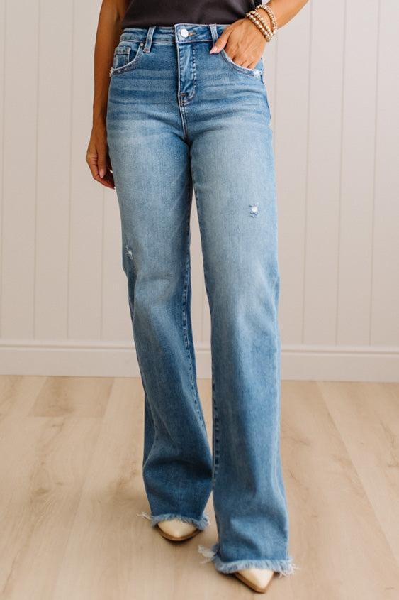 A person wearing light blue high-waisted flare jeans with a raw frayed hem, standing on a light wood floor against a white paneled wall.