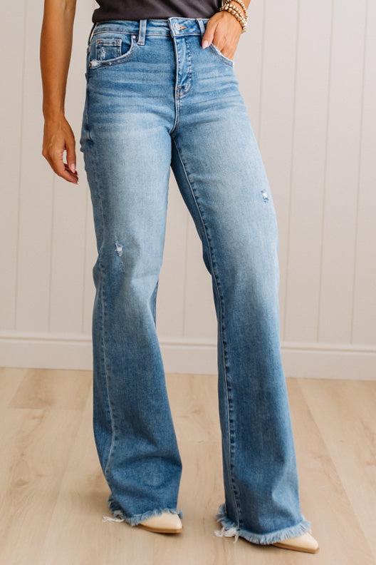 A person wearing light-wash blue flare jeans with distressed details and frayed hems, standing on a light wood floor against a white paneled wall.