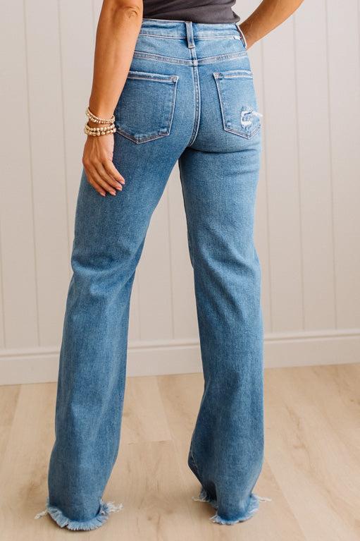 A person shown from the back wearing light blue wide-leg denim jeans with a raw frayed hem, standing on a light wood floor against a white paneled wall.