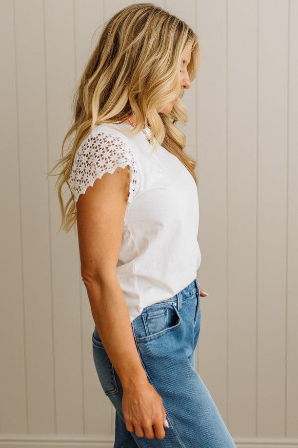 Woman wearing a white lace top and blue jeans against a beige wall.