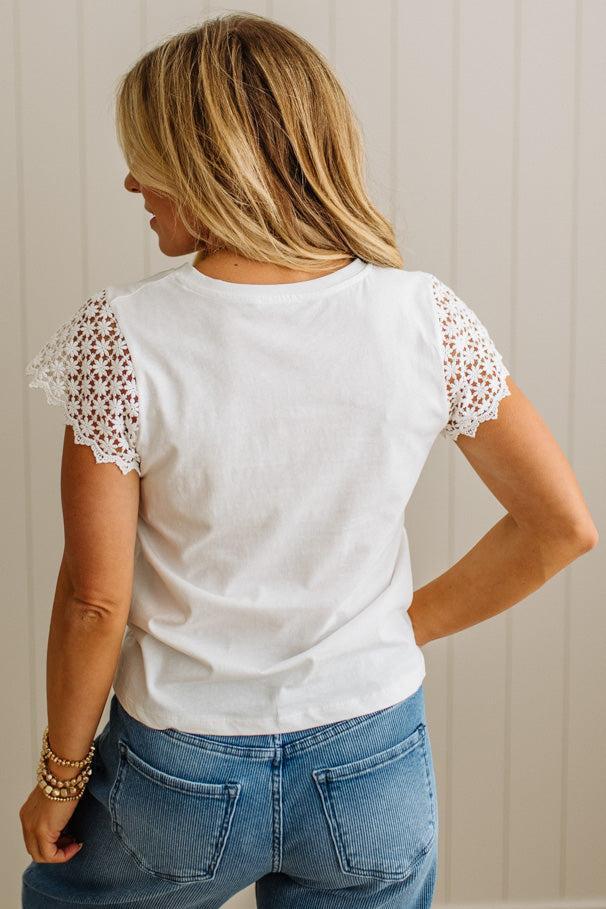 Person wearing a white t-shirt with lace sleeves and blue jeans against a neutral background