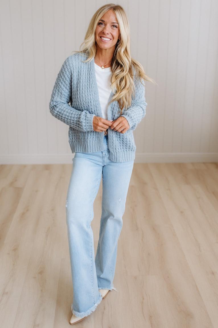 A woman wearing a light blue chunky cable-knit cardigan over a white top and light blue jeans, standing in a minimalist room with light wood floors and a beige wall.