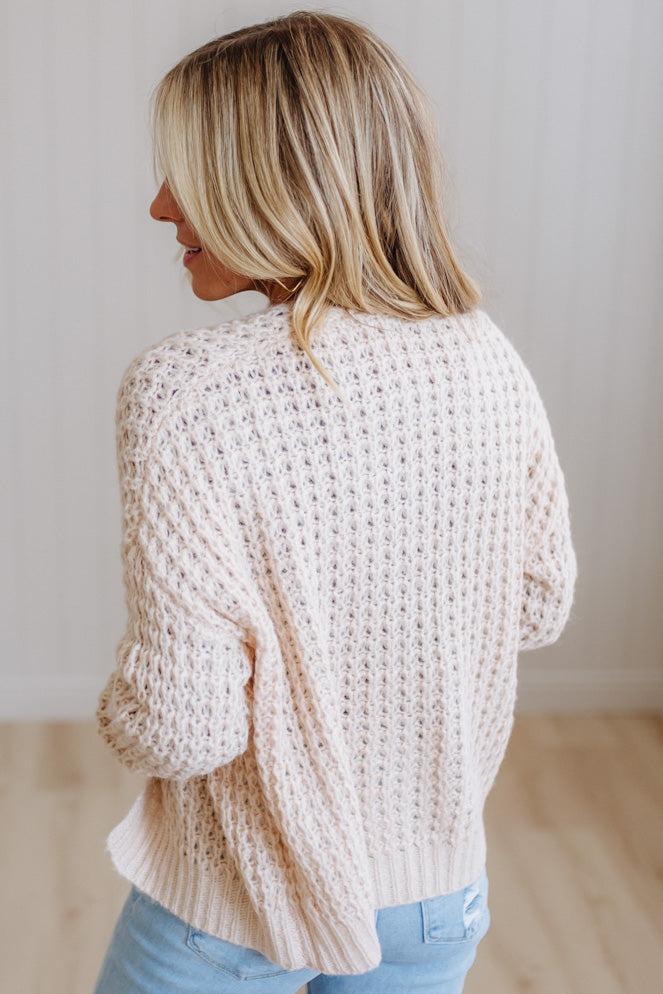 A woman with blonde hair is shown from the back wearing a cream-colored, chunky open-knit sweater and light blue jeans in a bright indoor setting.