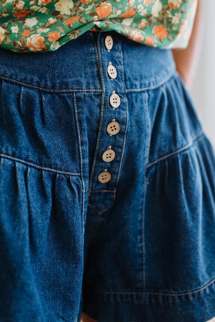 Woman in a green floral tank wearing dark wash denim shorts with wooden buttons at the front, and tiered flare cut.