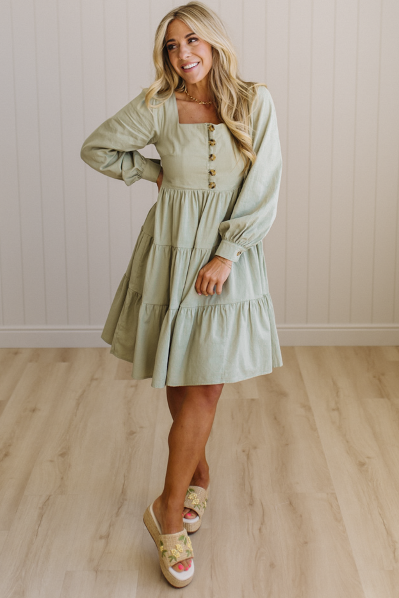 Woman wearing a light green dress with long sleeves in a room with wooden flooring and a white wall.