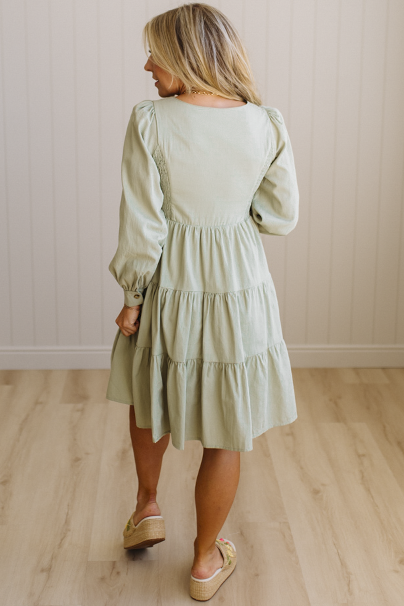 Woman wearing a light green dress with long sleeves in a room with wooden flooring and a white wall.