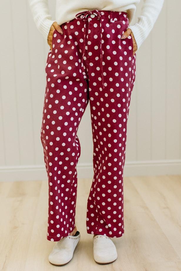 A person wearing red and white polka dot drawstring pants, a white sweater, and white slip-on shoes, standing in a room with light wood floors and white paneled walls.