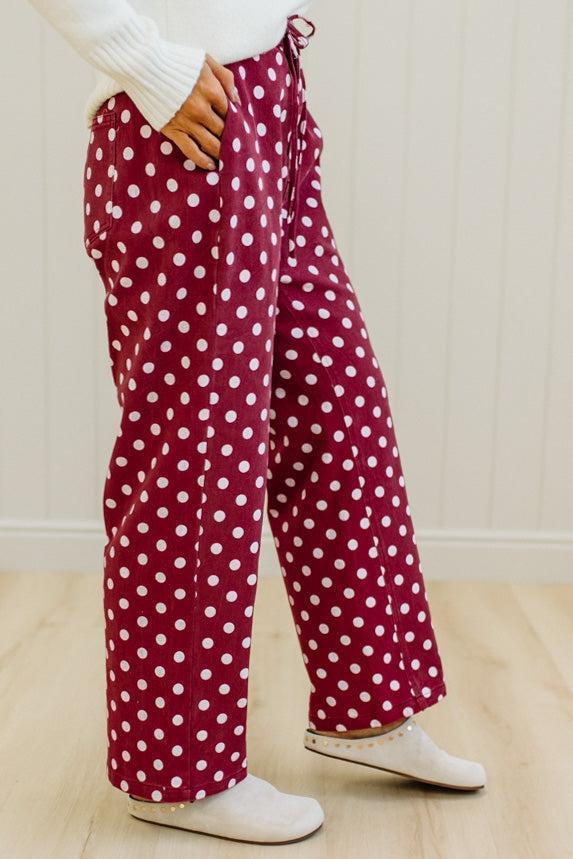 A person wearing burgundy wide-leg pajama pants with a white polka dot pattern, styled with a white ribbed knit top and white slip-on shoes.