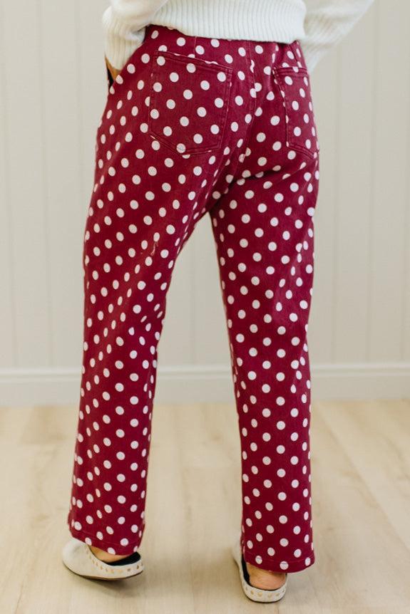 A person shown from the back wearing burgundy pants with a white polka dot pattern, paired with a white sweater and white slip-on shoes, standing on a light wood floor against a white paneled wall.