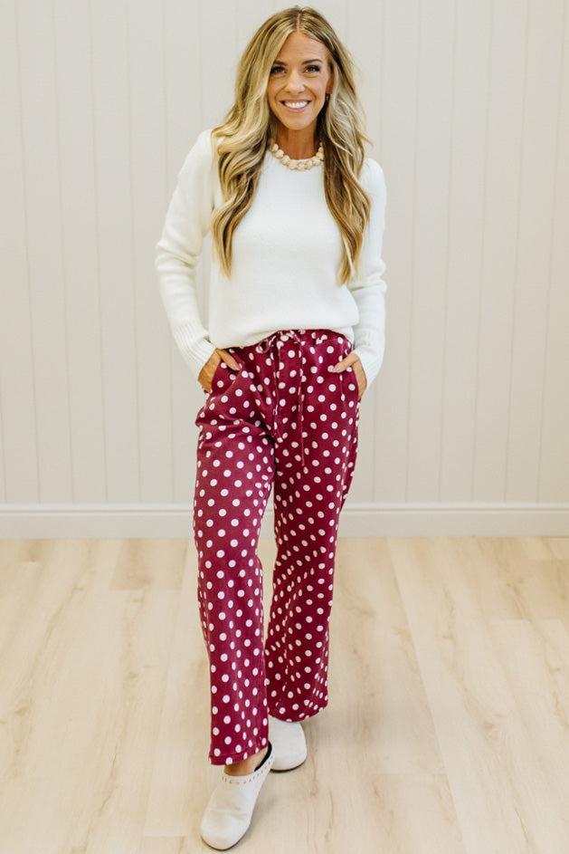 A woman modeling a white crewneck sweater and burgundy pants with white polka dots, standing on a light wood floor against a white paneled wall.