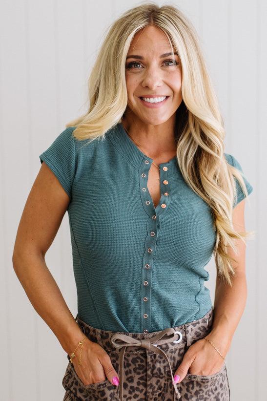 Woman wearing a teal short-sleeve top with buttons, standing against a plain background