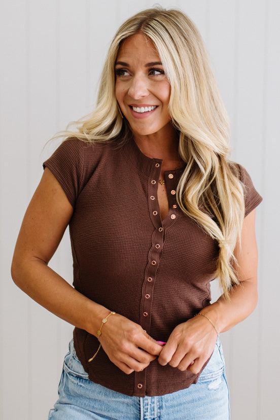 Woman wearing a brown short-sleeve top with a button-up front, standing against a plain background.