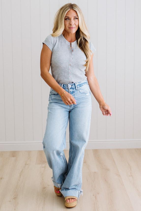 Woman wearing a gray top and light blue jeans standing against a white wall.