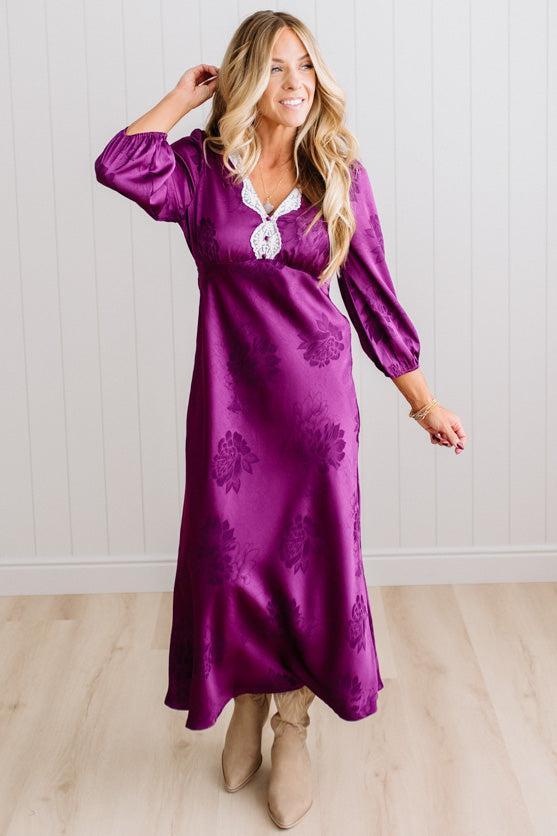 A woman modeling a plum-purple satin-like midi dress with a floral jacquard pattern, featuring a wrap-style collar and three-quarter sleeves, paired with white slide sandals against a white paneled wall and light wood floor.