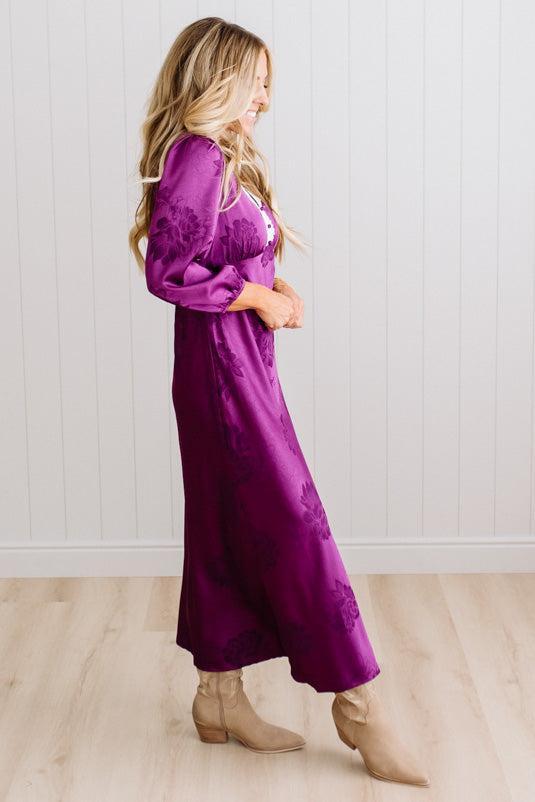A woman modeling a plum-purple satin-like midi dress with a floral jacquard pattern, featuring a wrap-style collar and three-quarter sleeves, paired with white slide sandals against a white paneled wall and light wood floor.
