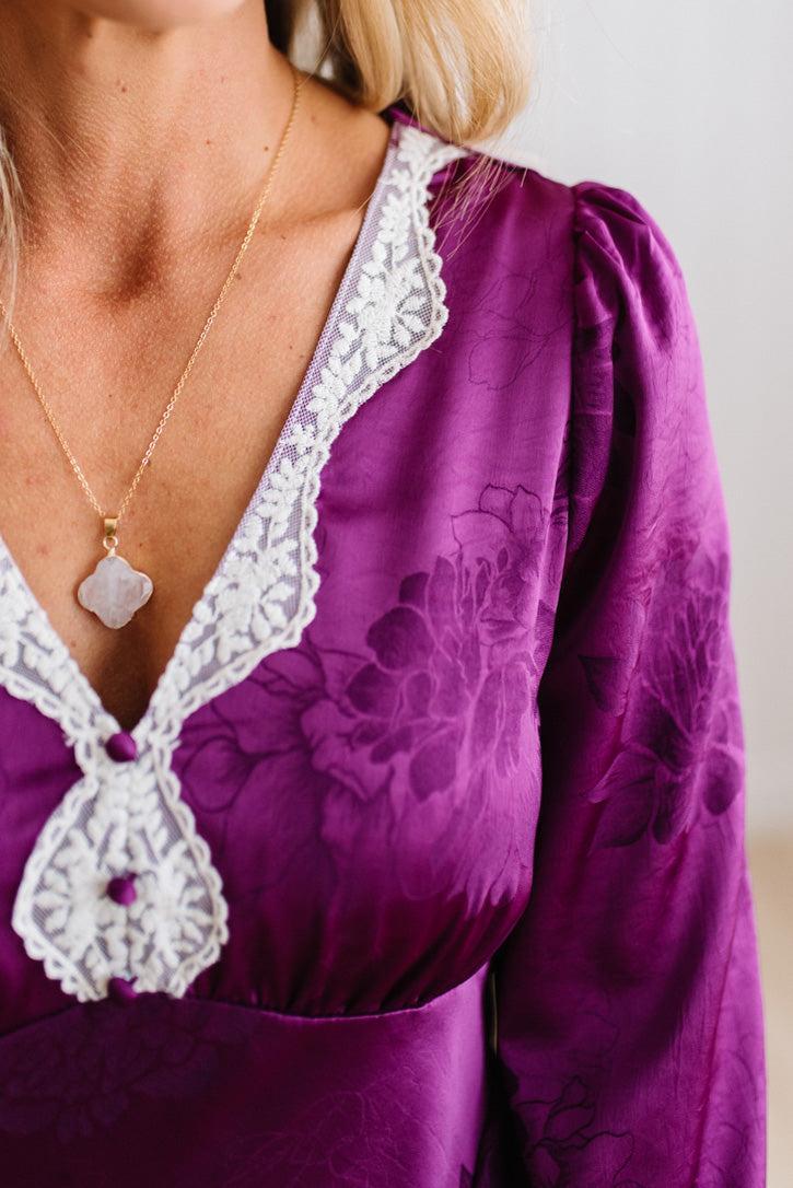 A woman modeling a plum-purple satin-like midi dress with a floral jacquard pattern, featuring a wrap-style collar and three-quarter sleeves, paired with white slide sandals against a white paneled wall and light wood floor.