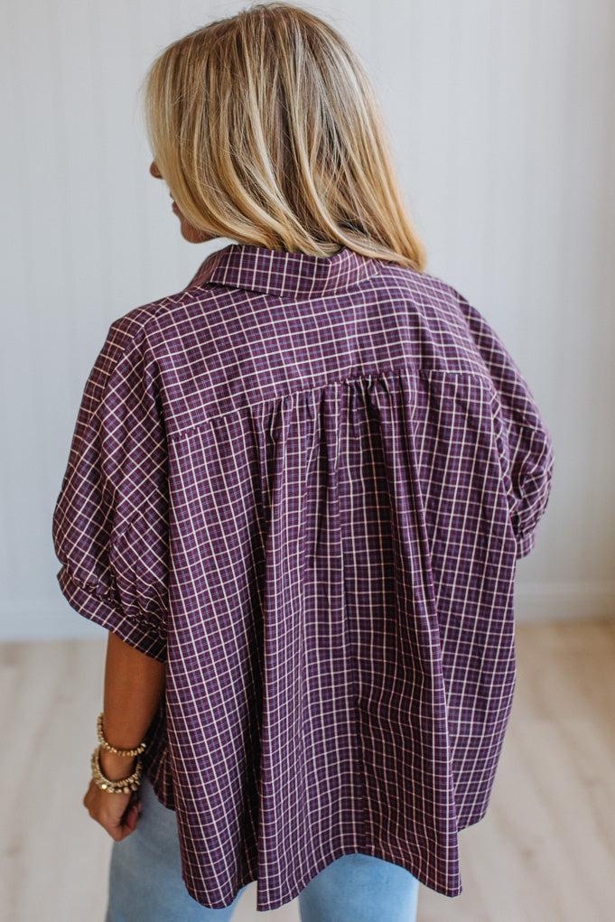A woman shown from the back wearing a loose, short-sleeve button-up blouse with a purple and white windowpane plaid pattern, paired with light blue jeans.