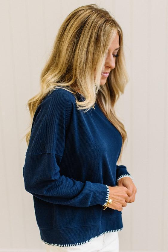 BLONDE WOMAN WEARING A WAFFLE TEXTURE TOP WITH A CREAM BLANKET STITCH TRIM, STANDING AGAINST A LIGHT WALL ON A LIGHT WOOD FLOOR. THE TOP IS NAVY.