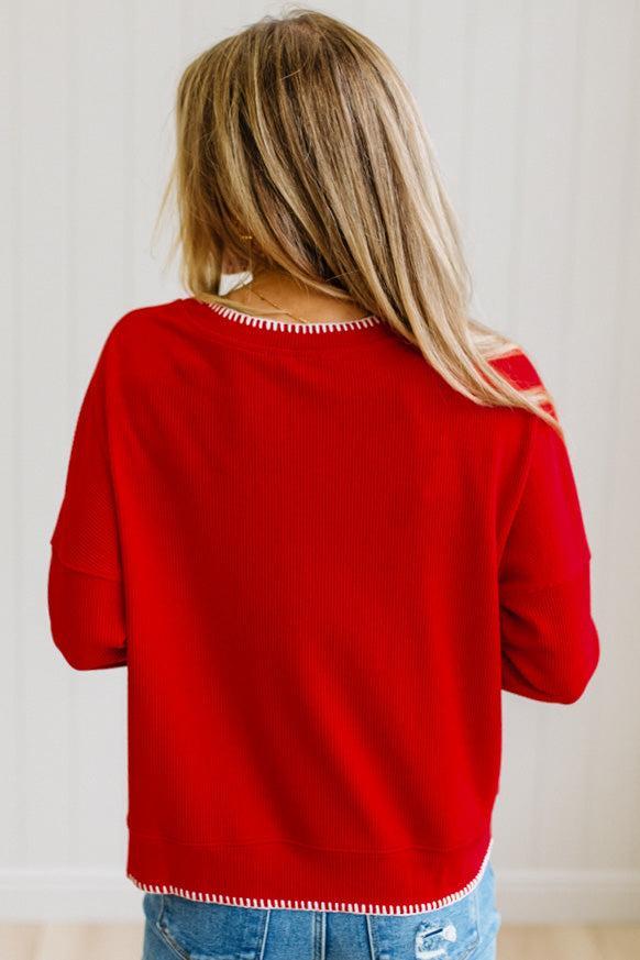 BLONDE WOMAN WEARING A WAFFLE TEXTURE TOP WITH A CREAM BLANKET STITCH TRIM, STANDING AGAINST A LIGHT WALL ON A LIGHT WOOD FLOOR. THE TOP IS RED.