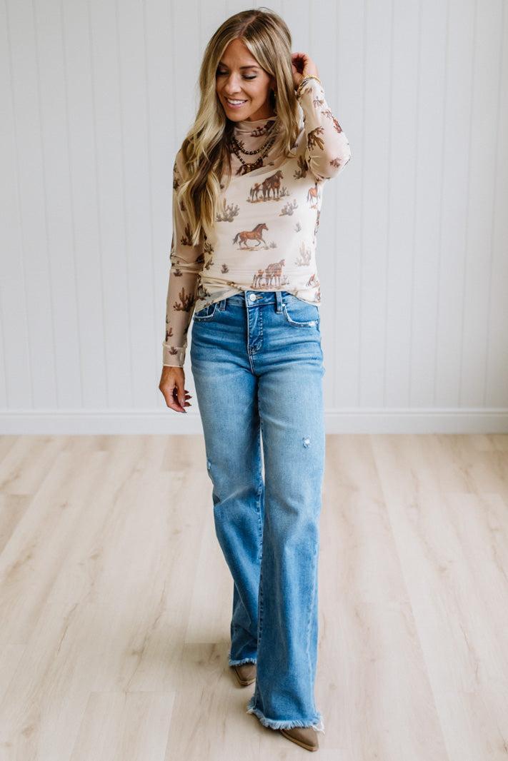 A woman wearing a beige long-sleeve turtleneck top with a western-themed horse print and blue flare jeans, standing in a room with light wood floors and white paneled walls.