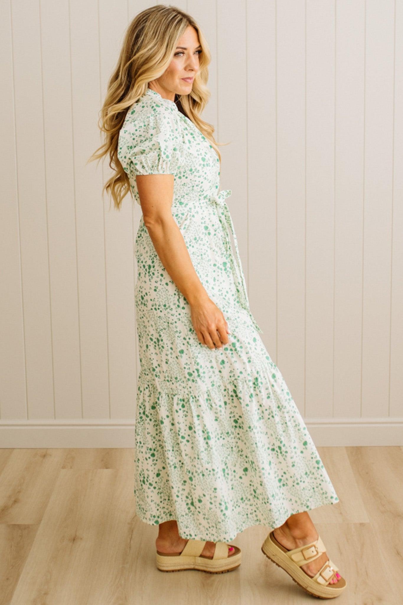 Woman wearing a floral dress standing against a wooden floor and white paneled wall.
