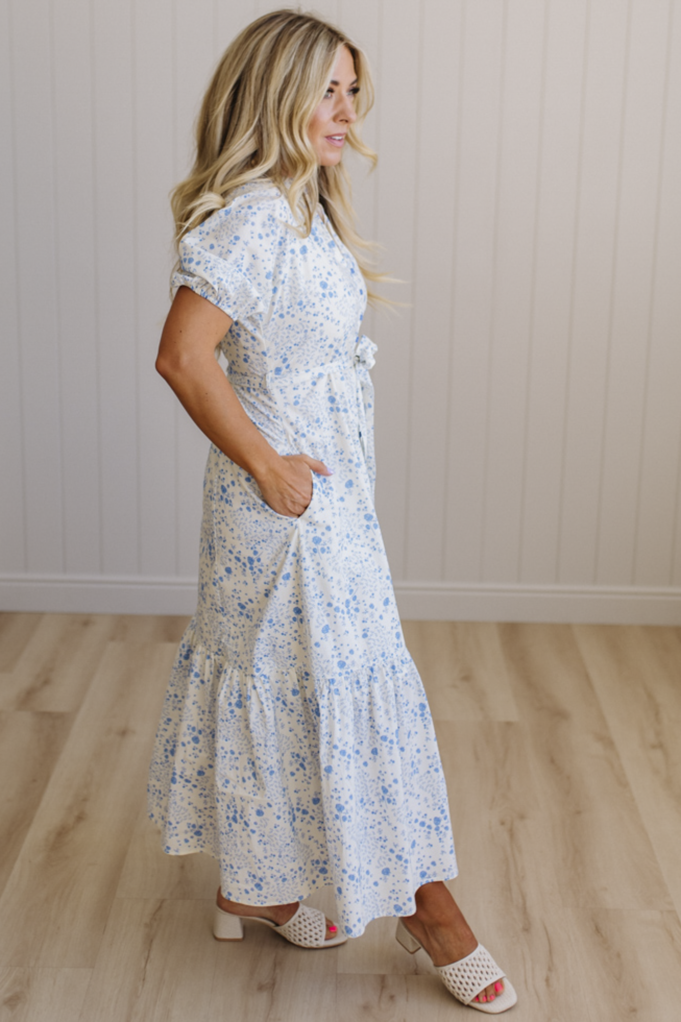 Woman wearing a floral dress standing on a wooden floor with a white paneled wall in the background