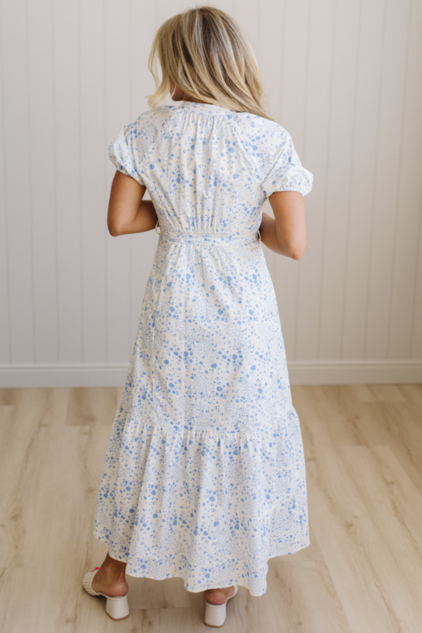 Woman wearing a white floral dress standing in a room with wooden flooring and a light-colored wall.