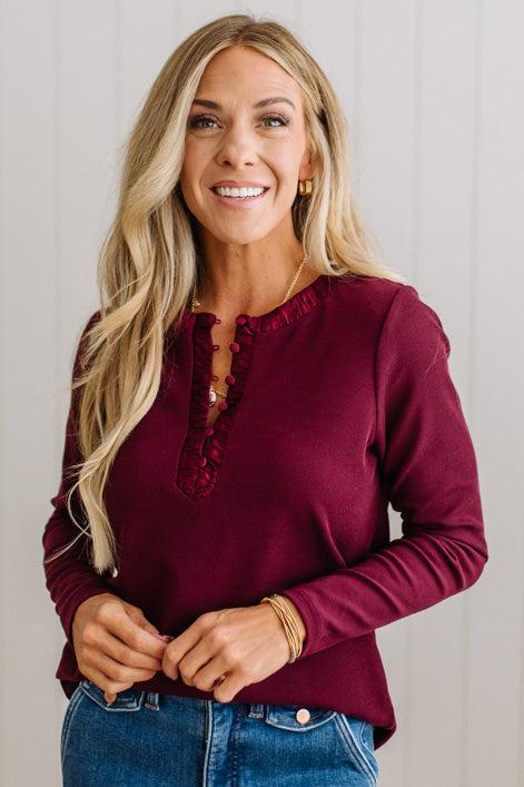 BLONDE WOMAN IN A BRIGHTLY LIT STUDIO WEARING DARK JEANS AND A SATIN RUCHED TRIM HALF BUTTON NECK, LONG SLEEVE, SOLID TOP IN BURGUNDY
