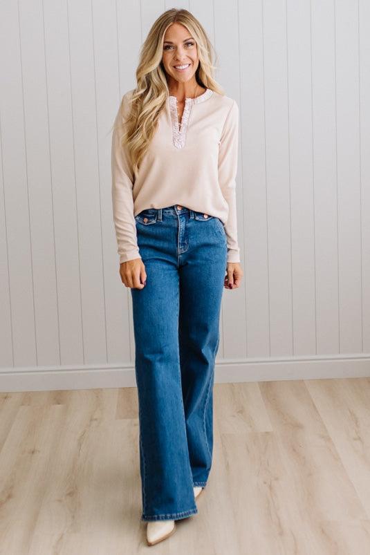 BLONDE WOMAN IN A BRIGHTLY LIT STUDIO WEARING DARK JEANS AND A SATIN RUCHED TRIM HALF BUTTON NECK, LONG SLEEVE, SOLID TOP IN IVORY