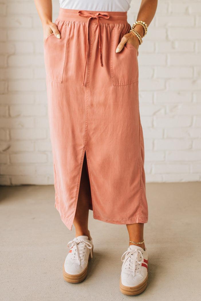Blonde woman in a hat wearing a white tank top and sneakers with a tencel cargo midi skirt with a center slit at the front.