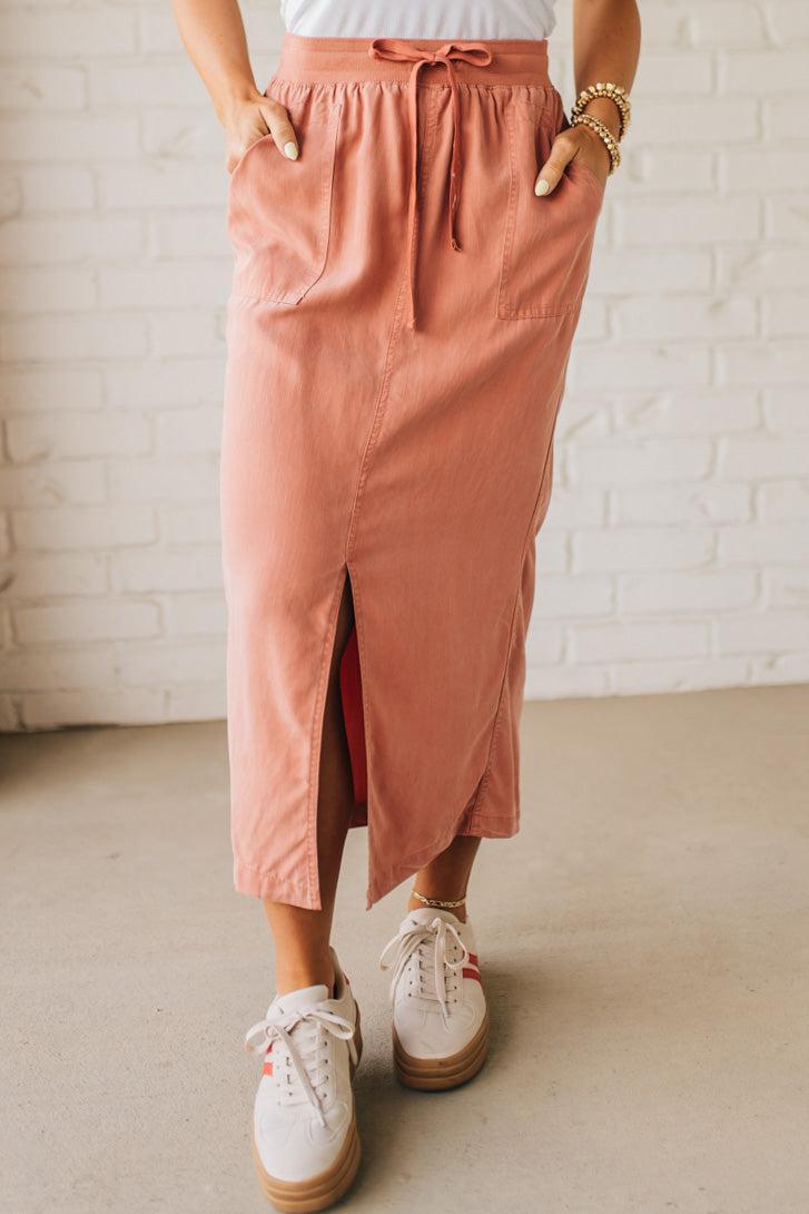 Blonde woman in a hat wearing a white tank top and sneakers with a tencel cargo midi skirt with a center slit at the front.