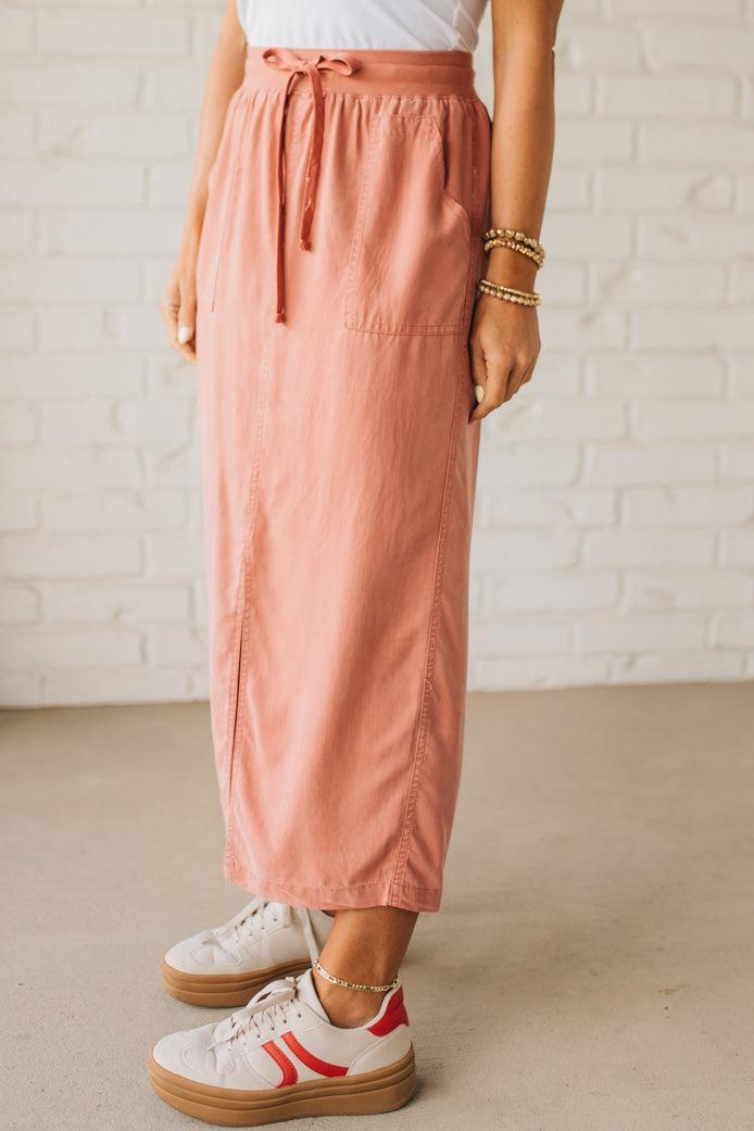 Blonde woman in a hat wearing a white tank top and sneakers with a tencel cargo midi skirt with a center slit at the front.