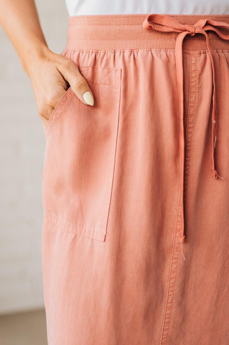 Blonde woman in a hat wearing a white tank top and sneakers with a tencel cargo midi skirt with a center slit at the front.