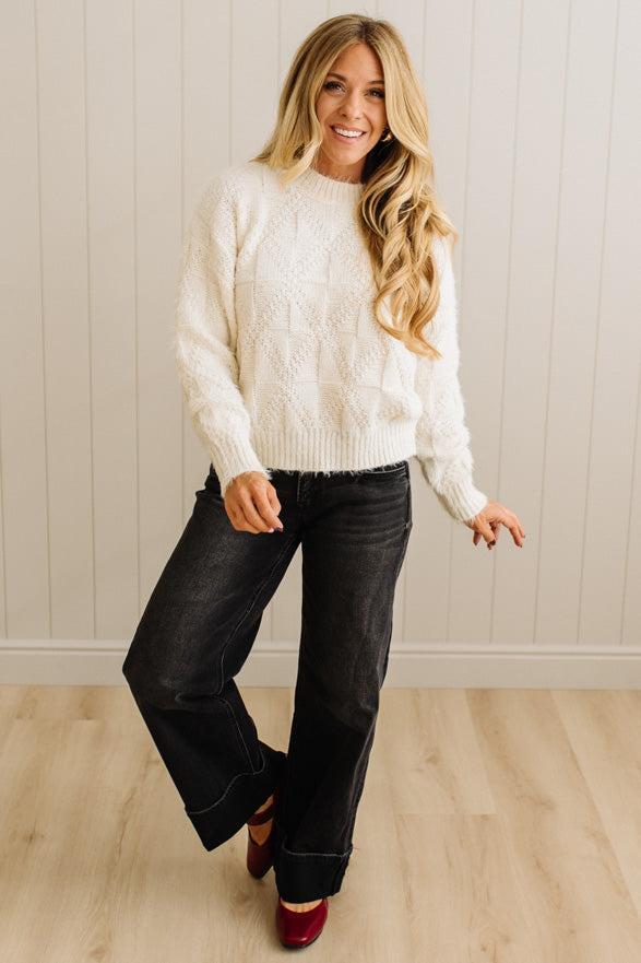 Woman wearing a white cable knit sweater and black jeans standing against a light wooden paneled wall.