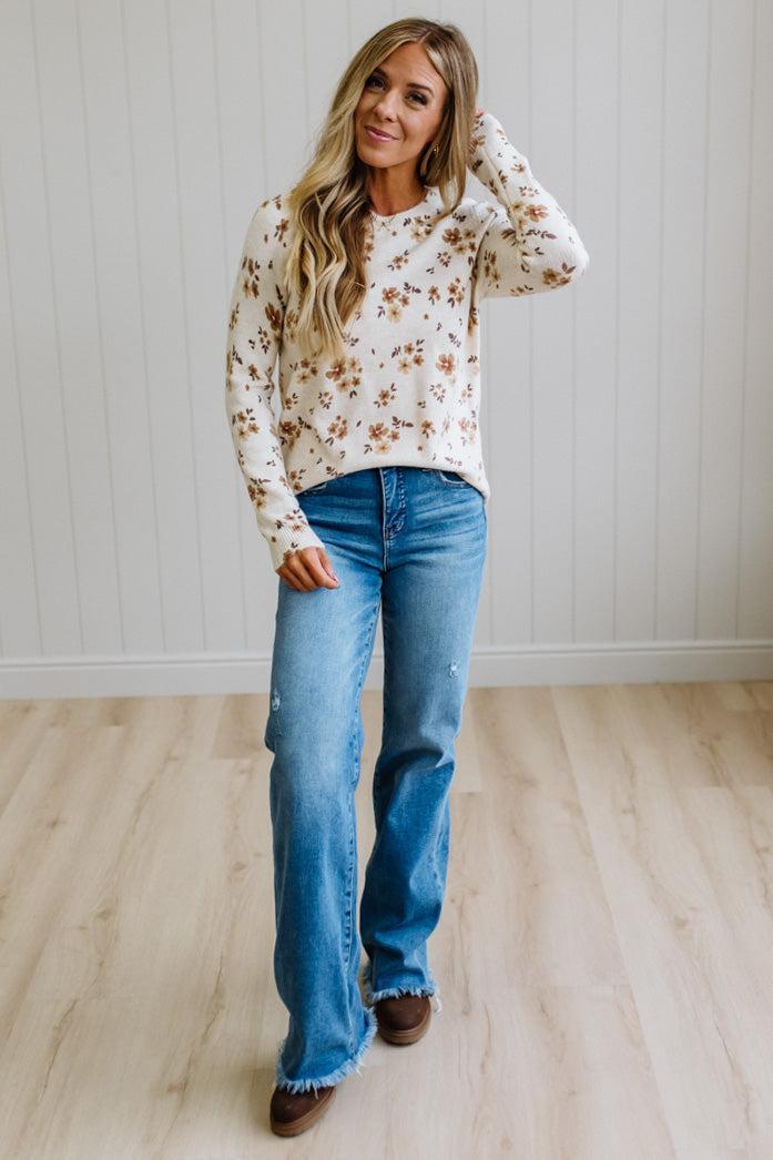 A woman modeling a cream-colored long-sleeve knit sweater with a brown floral pattern, paired with blue jeans and brown shoes, standing in a room with light wood floors and white paneled walls.