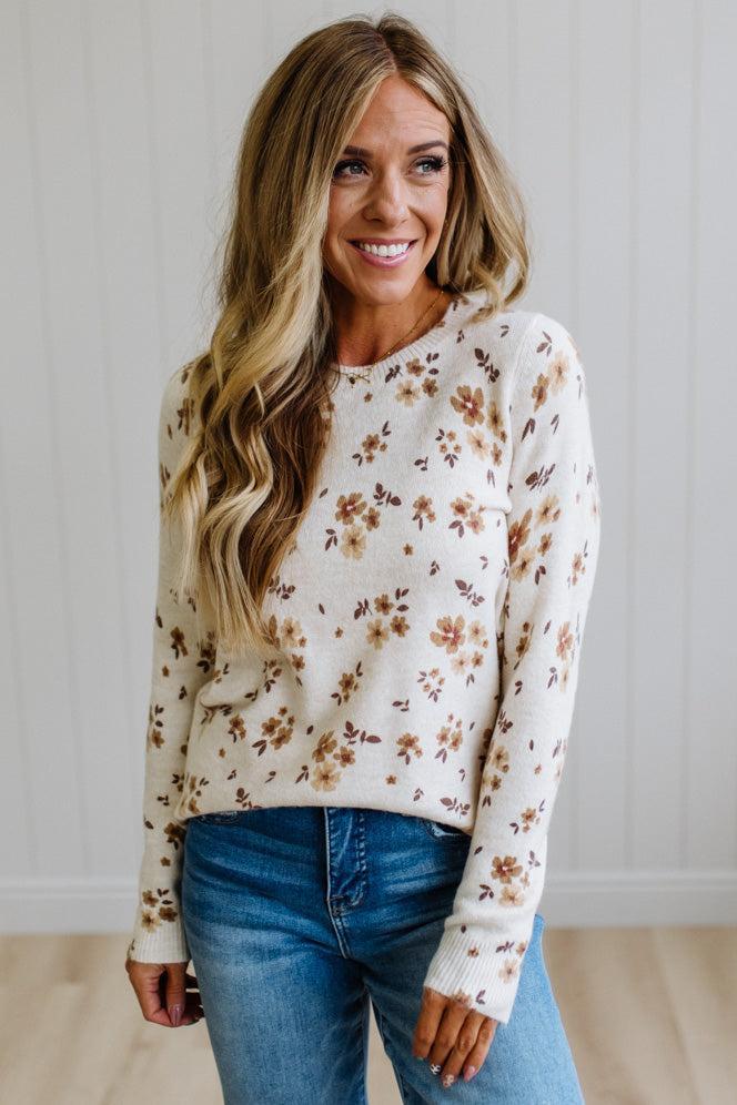 A woman with long blonde hair wearing a cream-colored knit sweater with a brown and tan floral pattern, paired with blue jeans, standing against a white paneled wall.